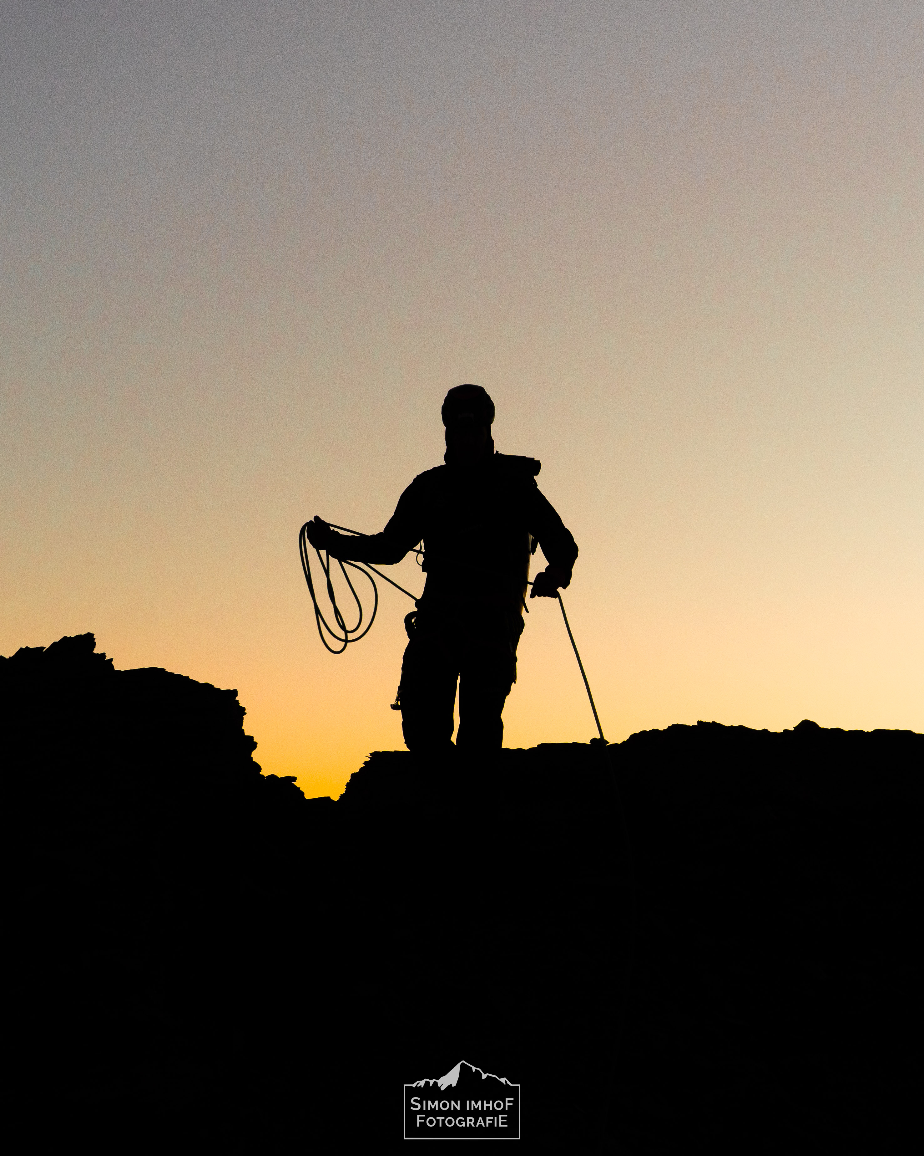 Sonnenaufgang. Die Silhouette des Bergführers zeichnet sich vor dem orangen Himmel ab., Seilhandling, Friends, Karabiner, Mensch auf Eiger Südgrat mit Fiescherhorn im Hintergrund, Adventures with Fabi, der Bergführer, Aussicht, Fabian Bendlin, Blick zurück auf dem Mittellegigrat, Überschreitung, Eiger Integral, Grat, Gratklettern, Bern, Simon Imhof, Alpen, Bergtour, Berner Oberland, Eiger, Gipfel, Gleitschirm, Hike and Fly, Hütte, Landschaftsfotografie, Mittellegigrat, Outdoor, Outdoorfotografie, SAC, Schweiz, Seilschaft, Simon Imhof, Sommer, Sonnenaufgang, Sony, Sportfotografie